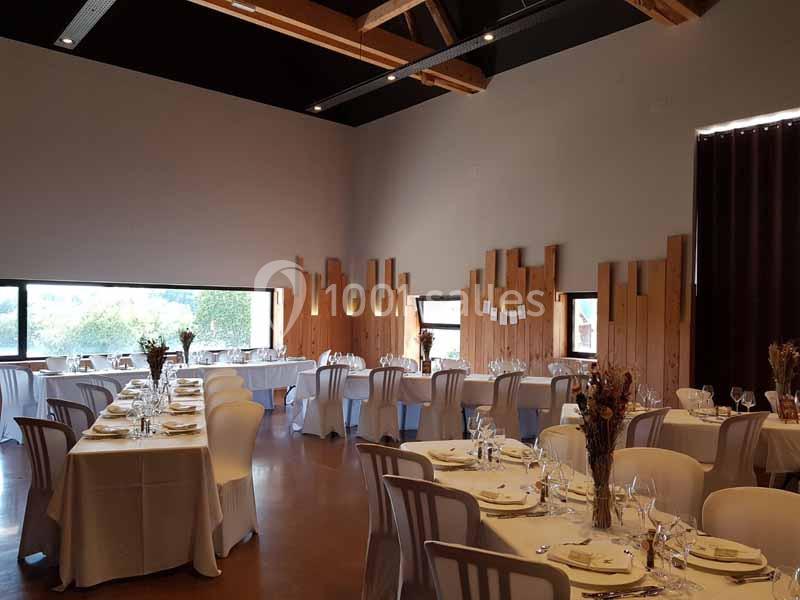 Salle de réception décorée avec des tables rondes dressées, chaises blanches et murs en bois clair.