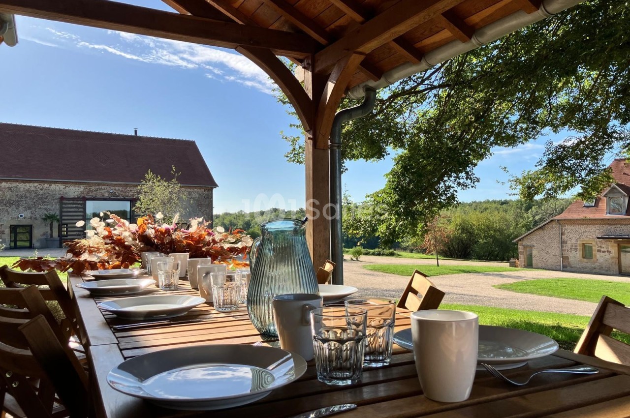 Table en bois dressée avec vaisselle et carafe, sous une pergola, donnant sur un jardin et des bâtiments ruraux.