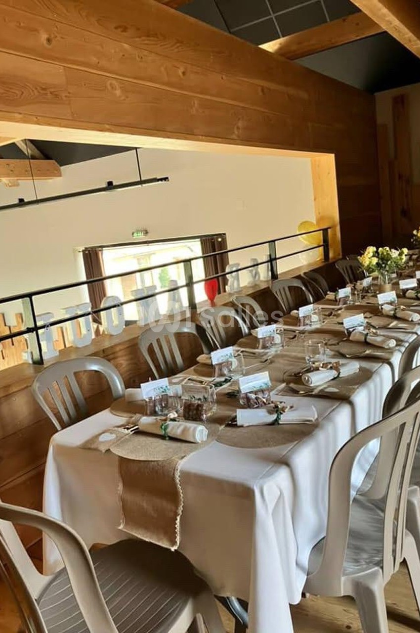 Table dressée avec nappes blanches, couverts, décorations rustiques et chaises dans une salle en bois lumineuse.