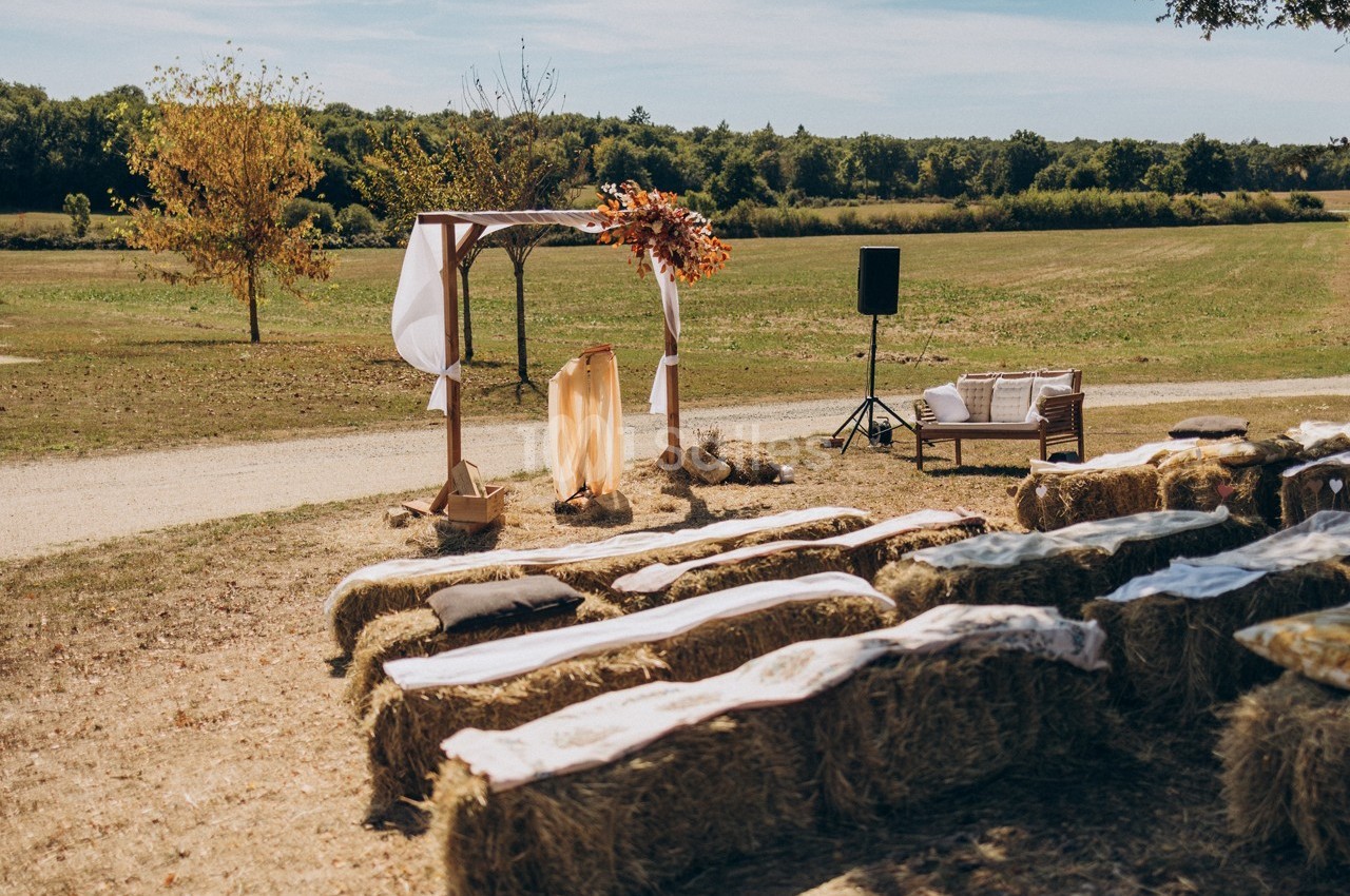 Cérémonie en plein air avec des bottes de foin en guise de sièges et une arche décorée de fleurs dans un champ.