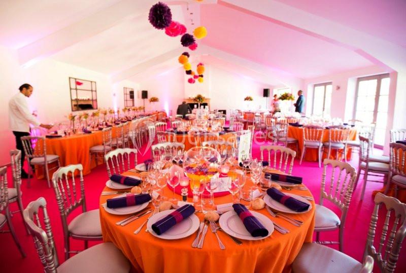 Salle de réception décorée avec des tables rondes orange, nappes colorées, et lumières roses.