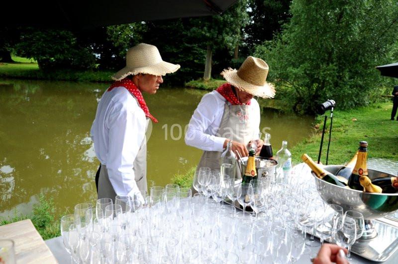 Deux personnes en tabliers et chapeaux de paille préparent des boissons près d'une table garnie de verres et de champagne.