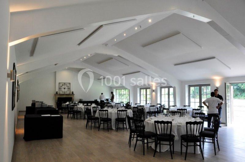 Salle lumineuse avec tables rondes dressées, chaises noires et grandes fenêtres donnant sur un espace extérieur verdoyant.