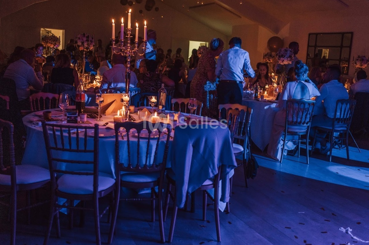 Salle de réception éclairée par des bougies, avec des tables décorées et des invités assis ou debout.