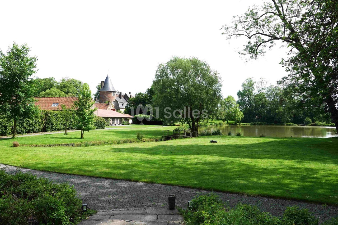 Vue d'un jardin verdoyant avec pelouse, arbres, un étang et une bâtisse en arrière-plan surmontée d'une tour.