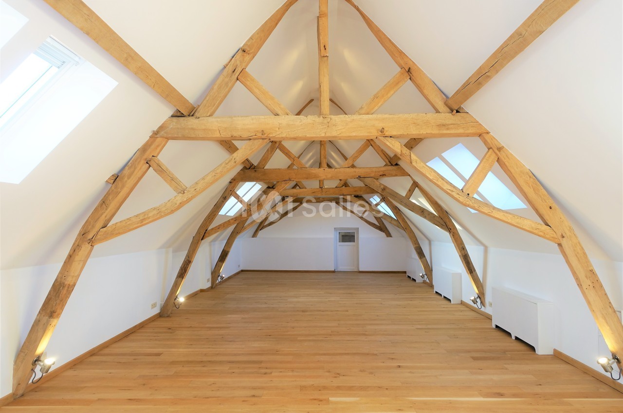 Combles aménagés avec poutres apparentes en bois, parquet clair et lumière naturelle provenant de fenêtres de toit.