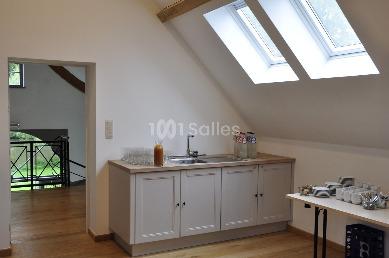 Petite kitchenette lumineuse avec évier, bouteilles d'eau, verres et vaisselle, sous un toit en pente avec fenêtres.