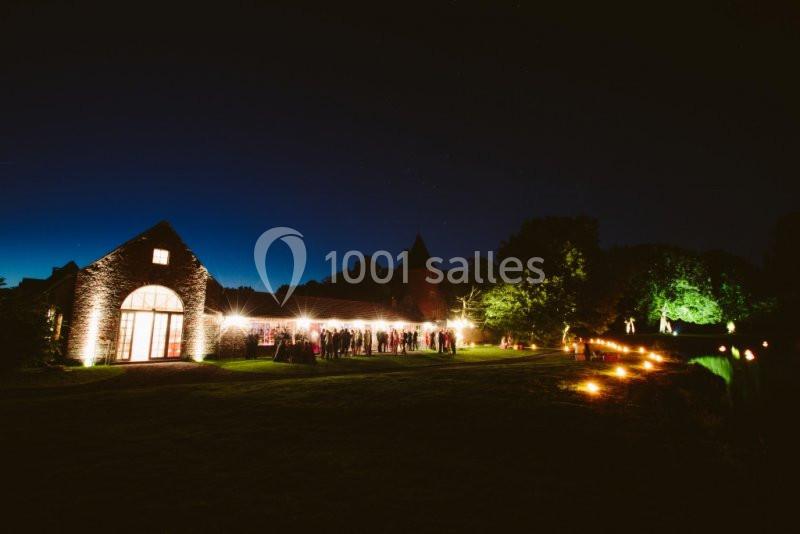 Bâtiment en pierre éclairé la nuit, entouré de lumières et de verdure, avec des silhouettes de personnes à l'extérieur.