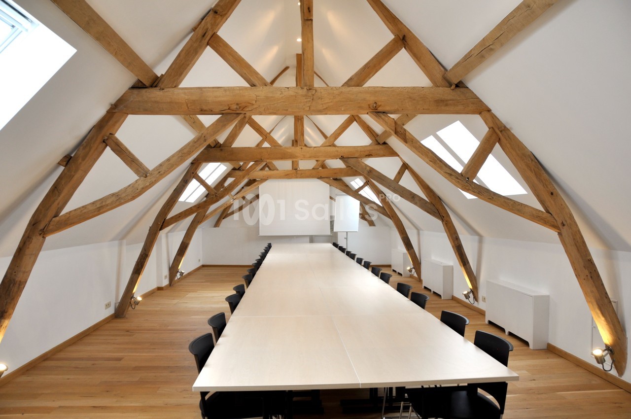 Salle de réunion spacieuse sous des poutres apparentes en bois, avec une grande table et des chaises noires.