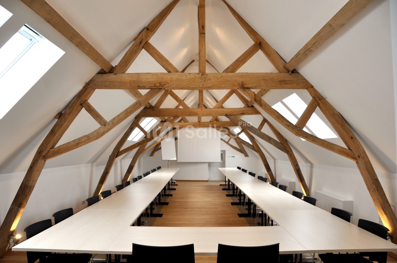 Salle de réunion lumineuse sous une charpente en bois apparente, équipée de longues tables et de chaises noires.