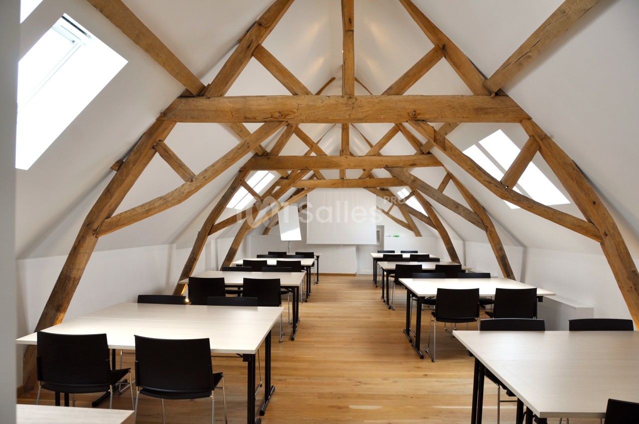 Salle lumineuse sous combles avec poutres apparentes, équipée de tables et chaises alignées pour travail ou réunion.