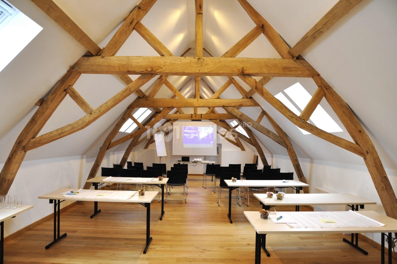 Salle de réunion lumineuse sous combles avec poutres apparentes, tables disposées en U et écran de projection.