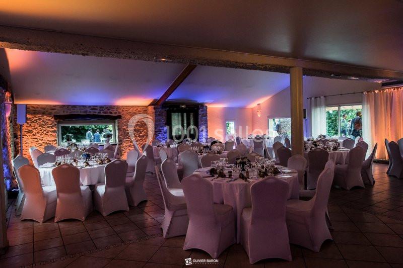 Salle de réception décorée avec des tables rondes dressées, chaises blanches et éclairage tamisé coloré.