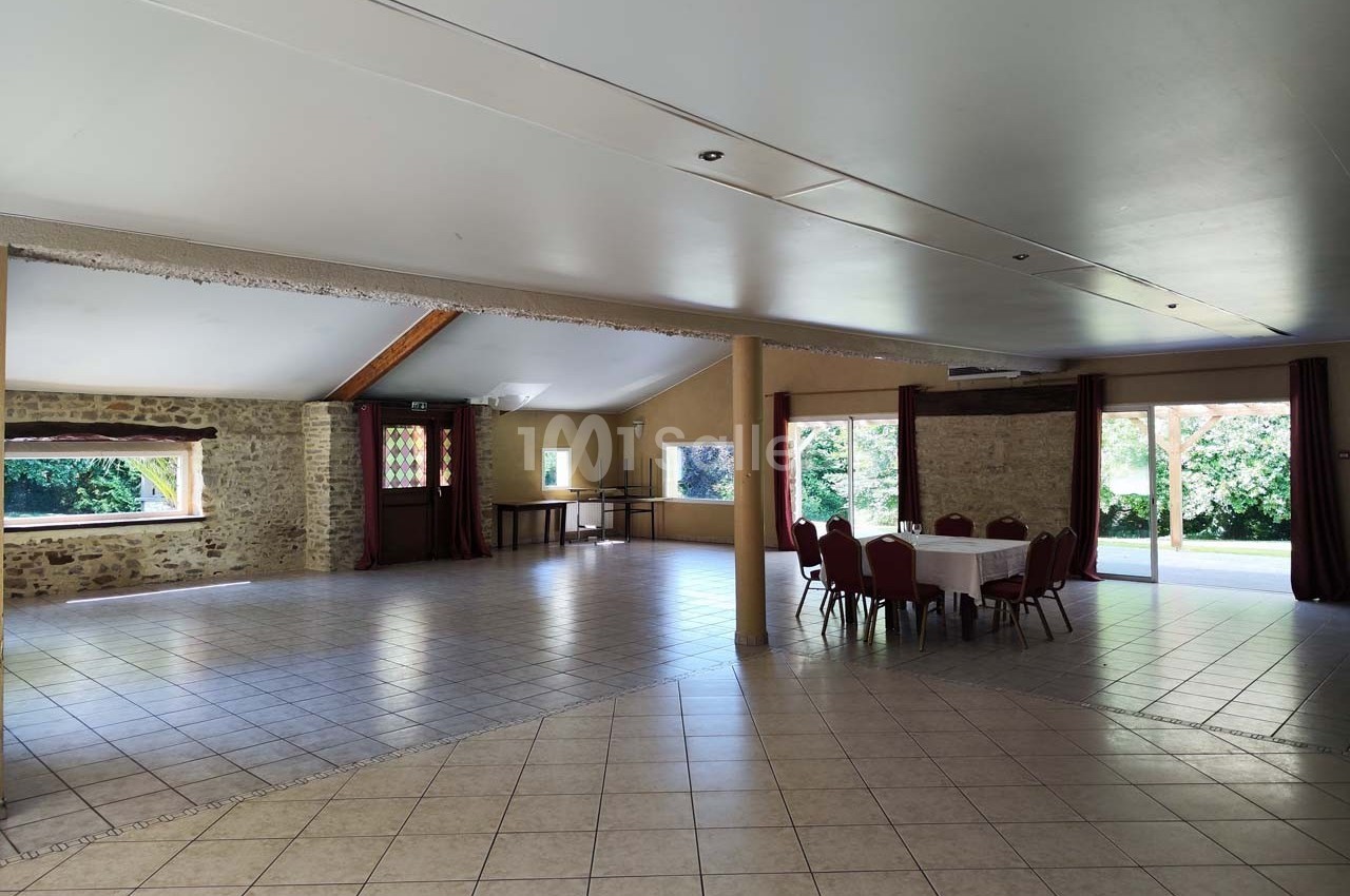 Salle spacieuse avec murs en pierre, grandes fenêtres, carrelage clair et quelques tables et chaises disposées.
