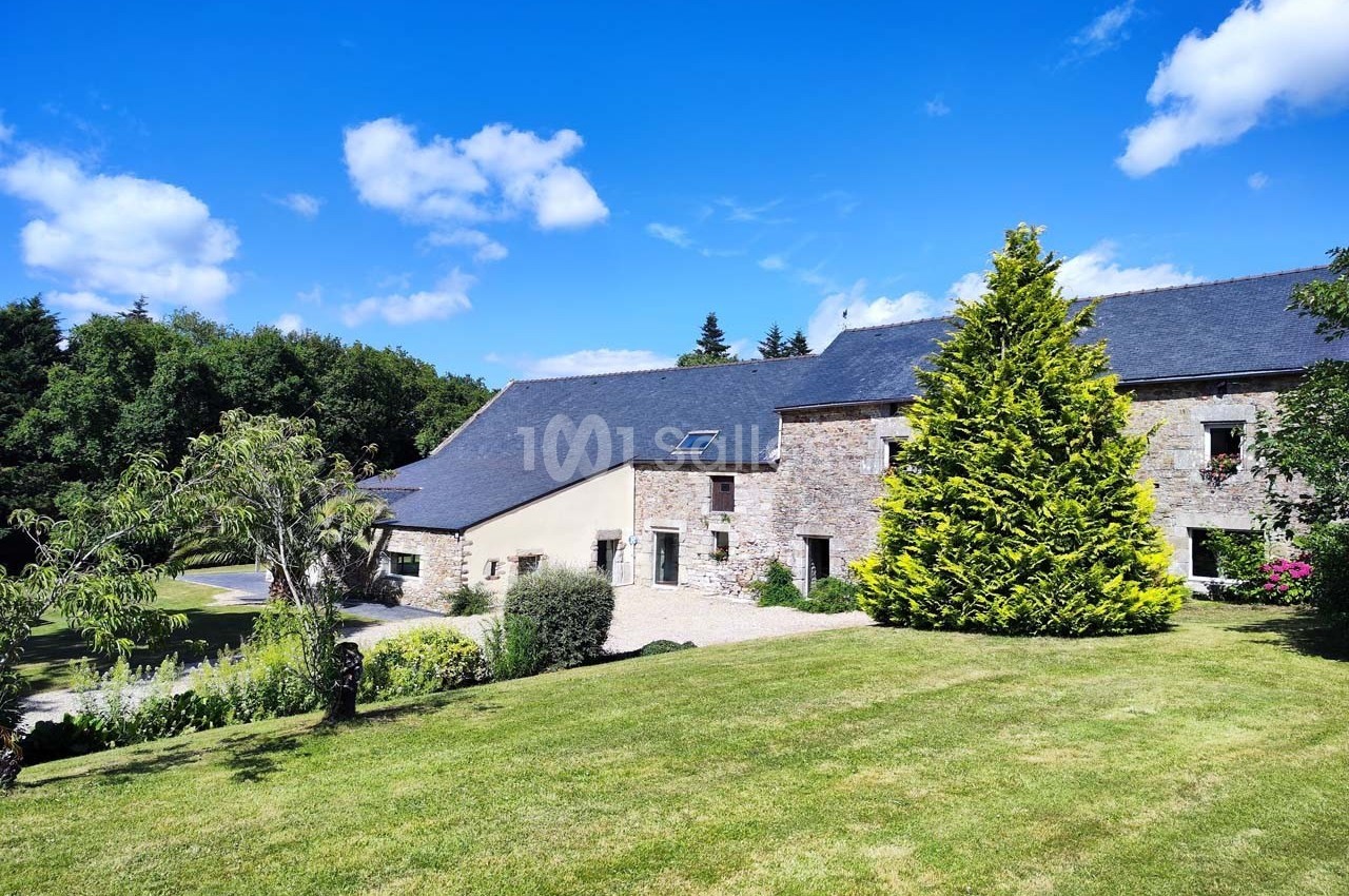 Bâtisse en pierre avec toit en ardoise entourée de verdure et d'un jardin sous un ciel bleu.