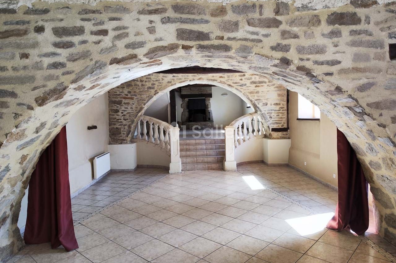 Salle voûtée en pierre avec sol carrelé, escaliers menant à une cheminée et rideaux rouges encadrant les ouvertures.