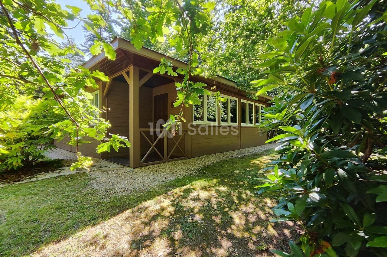 Chalet en bois entouré de végétation dense, avec un chemin de gravier à l'ombre des arbres.