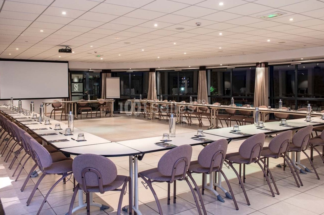 Salle de réunion spacieuse avec tables disposées en U, chaises alignées et écran de projection en fond.