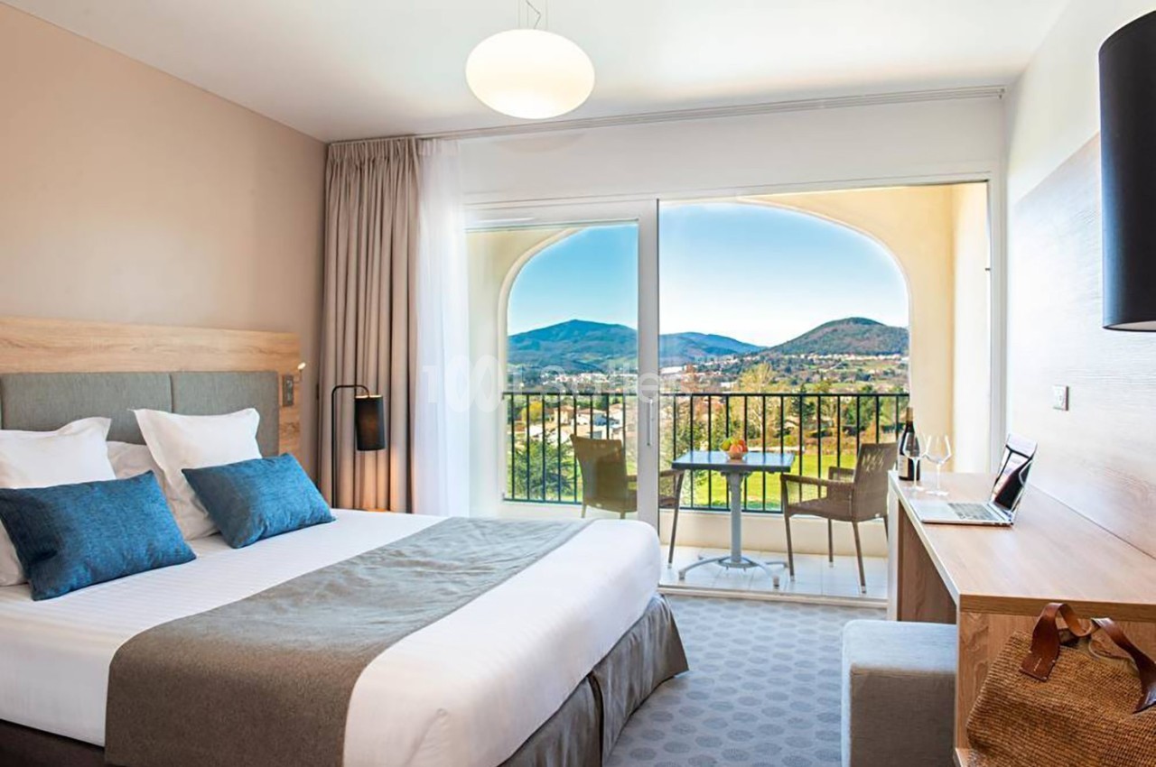Chambre lumineuse avec lit double, bureau, balcon et vue sur des collines verdoyantes à travers une baie vitrée.