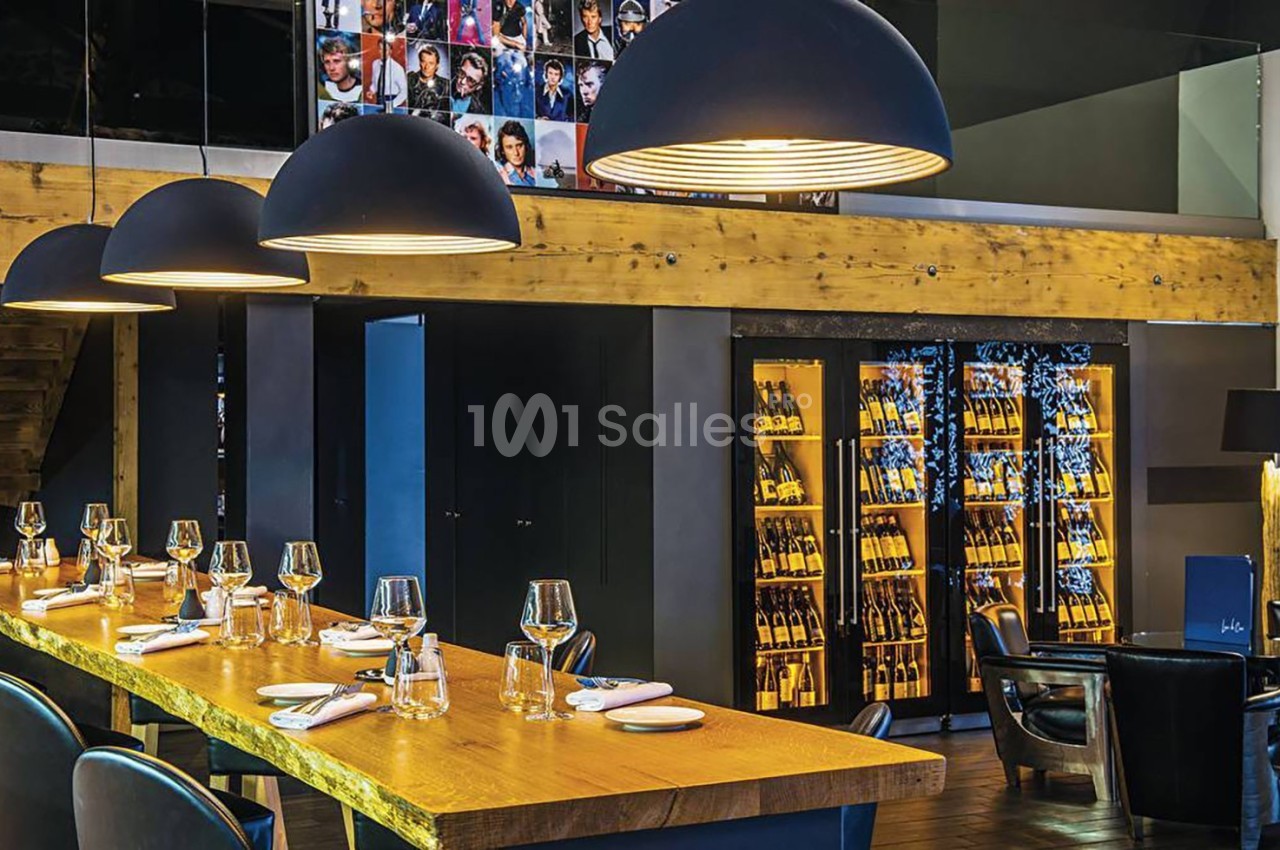 Salle de restaurant moderne avec une grande table en bois, suspensions noires et étagères de bouteilles illuminées.