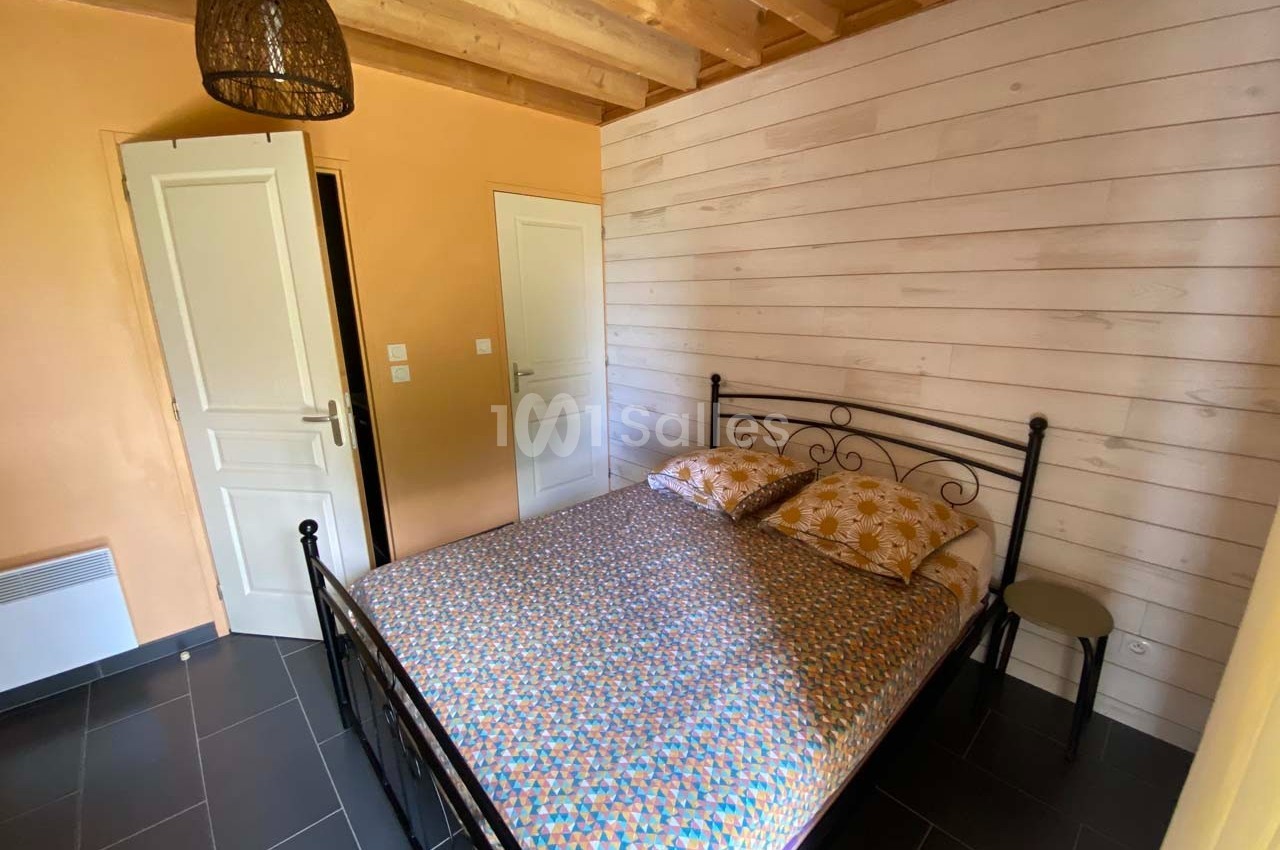 Chambre avec lit double en métal, murs en bois clair et orange, sol en carrelage foncé et plafond avec poutres apparentes.