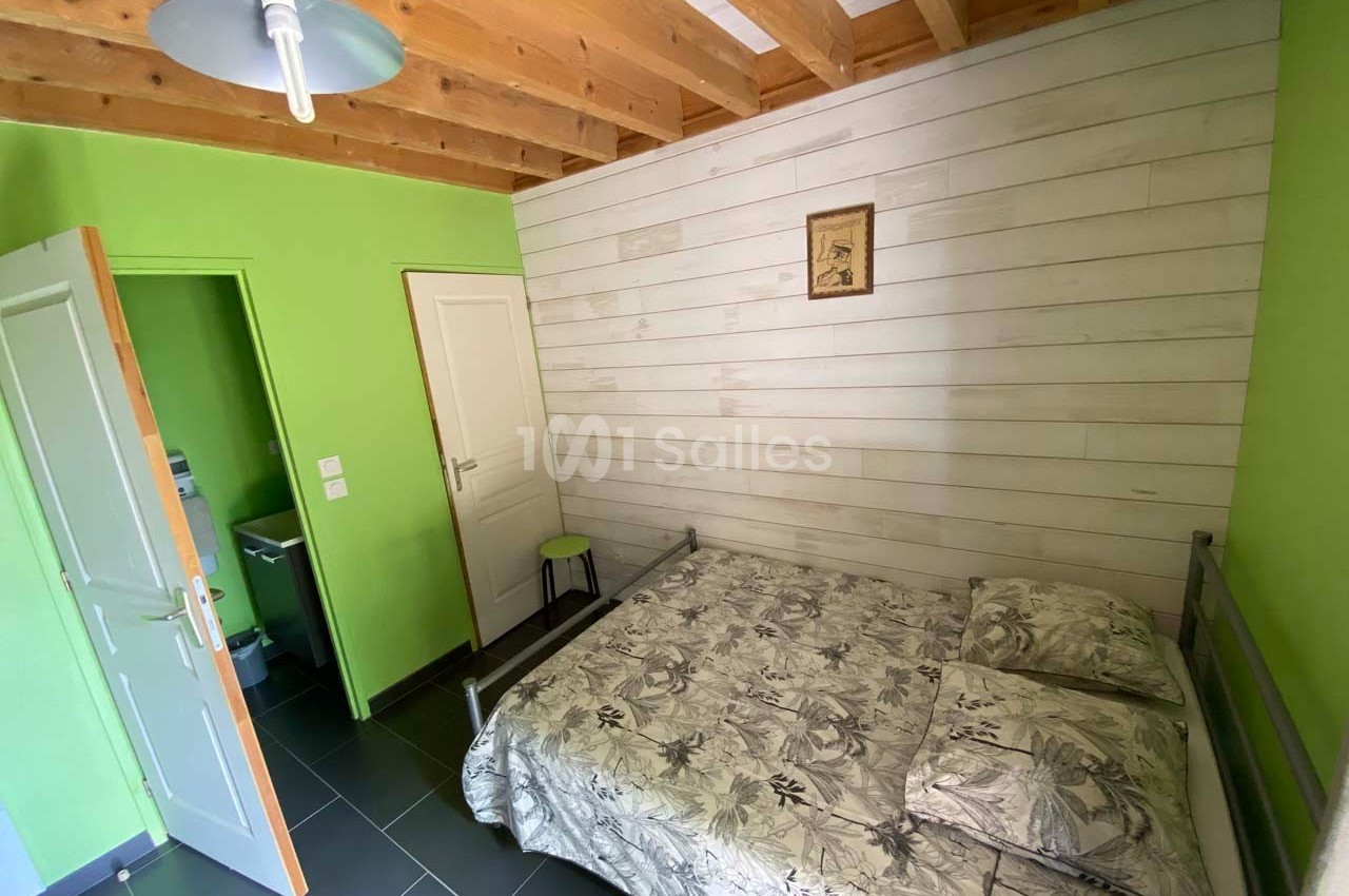 Chambre avec lit double, murs verts et en bois clair, plafond avec poutres apparentes, porte ouverte sur une salle d'eau.