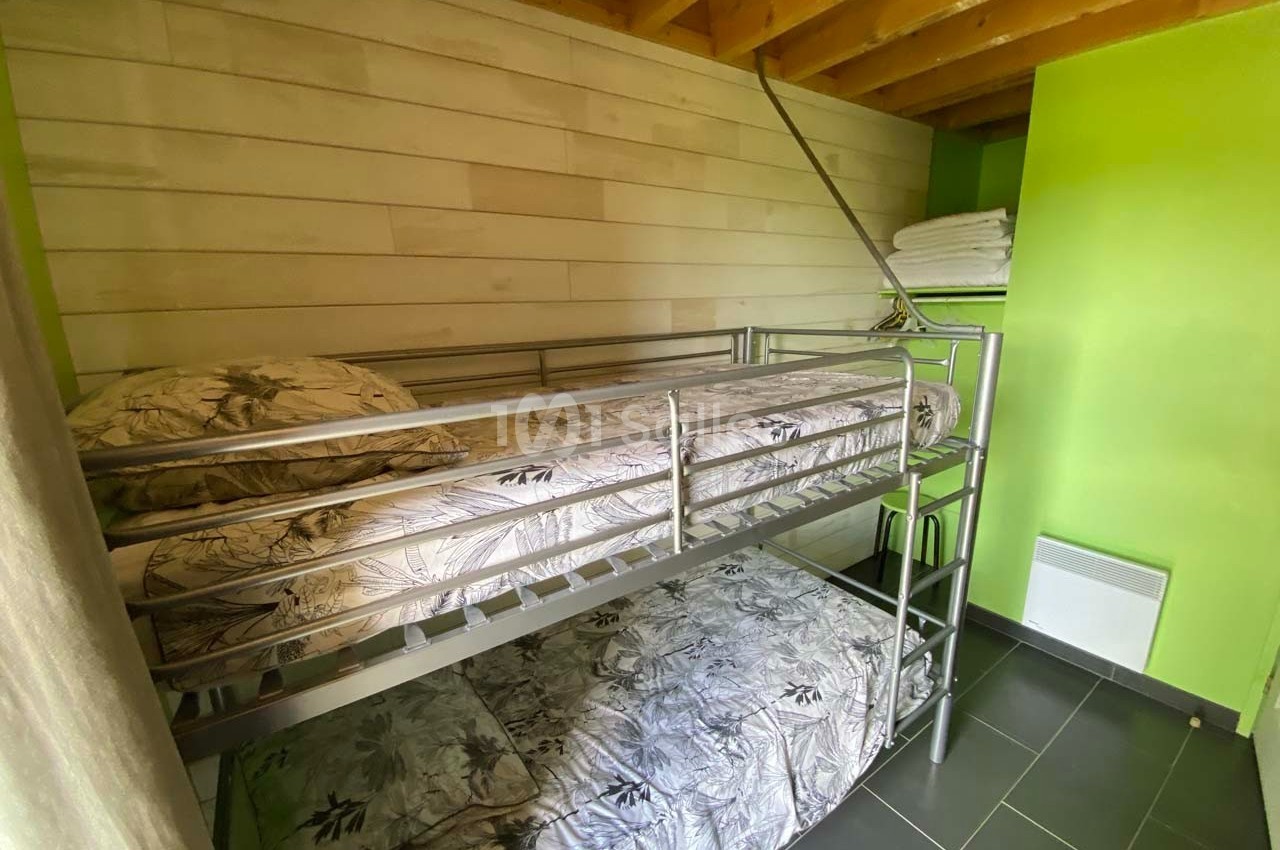 Lit superposé en métal dans une chambre aux murs verts et bois, avec des draps à motifs floraux et des serviettes pliées.