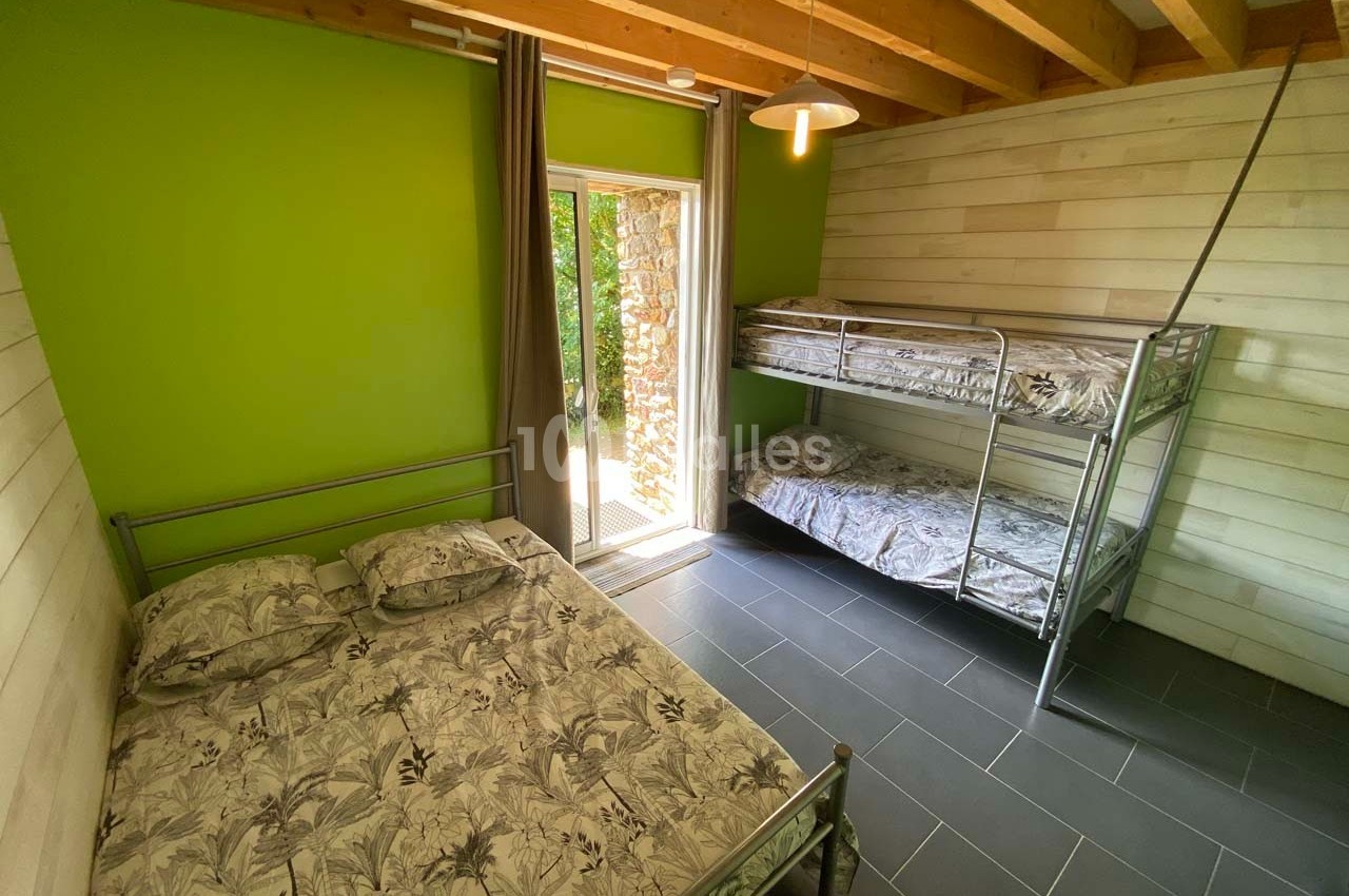 Chambre avec murs en bois et vert, lit double, lits superposés en métal, sol en carrelage gris et porte-fenêtre donnant sur…