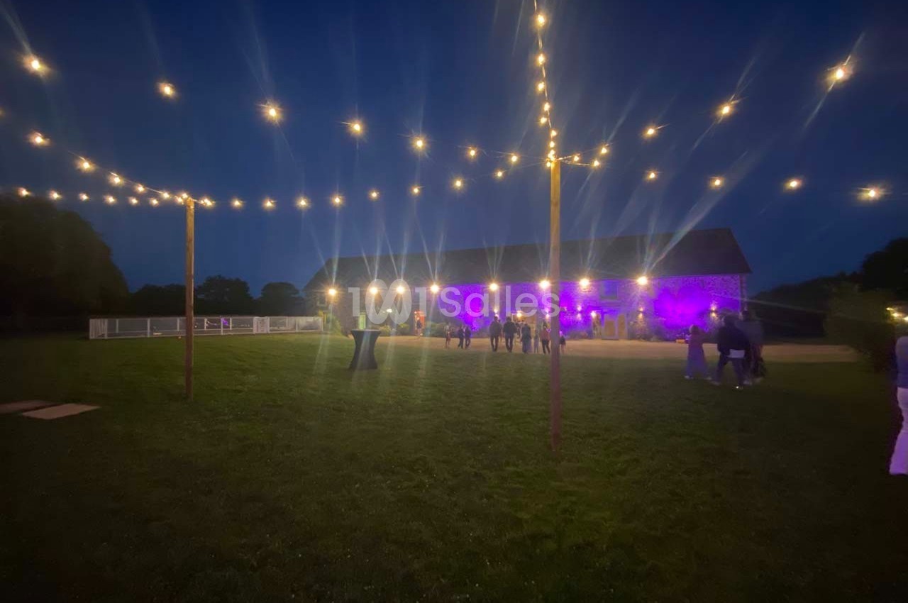 Guirlandes lumineuses suspendues au-dessus d'un espace extérieur sombre avec une grange éclairée en arrière-plan.
