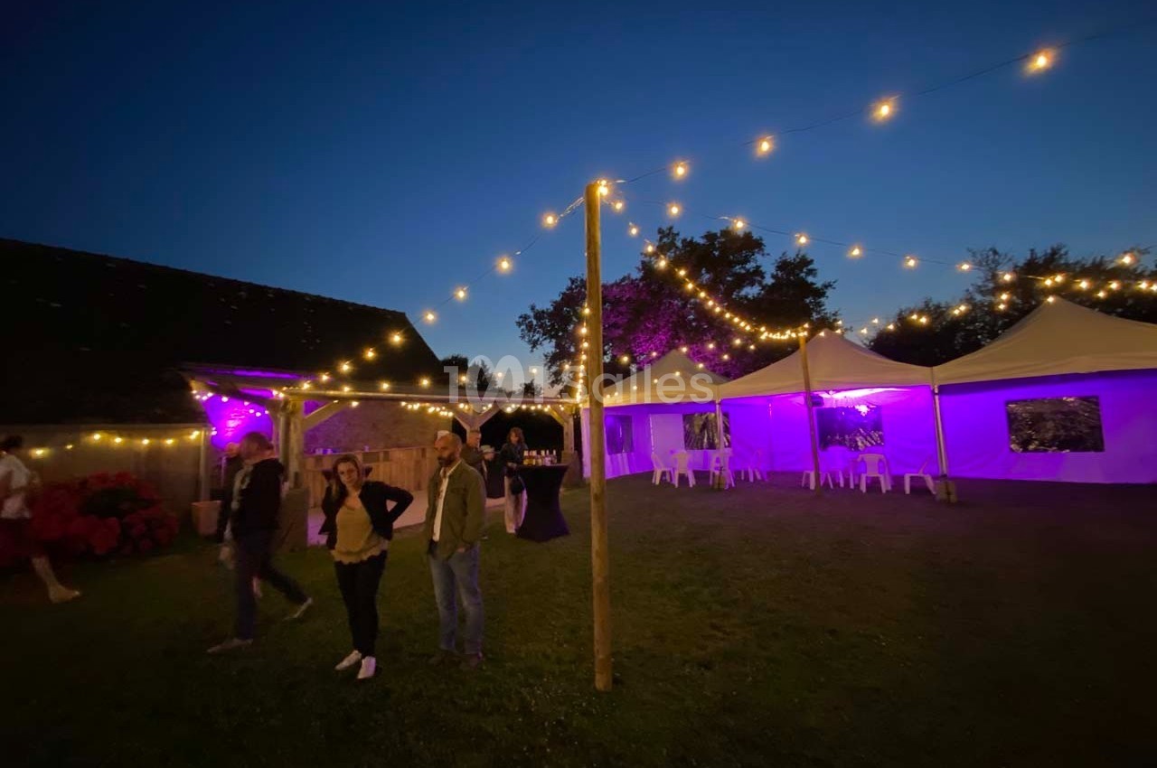 Tentes éclairées en violet et guirlandes lumineuses dans un jardin au crépuscule, avec quelques personnes présentes.