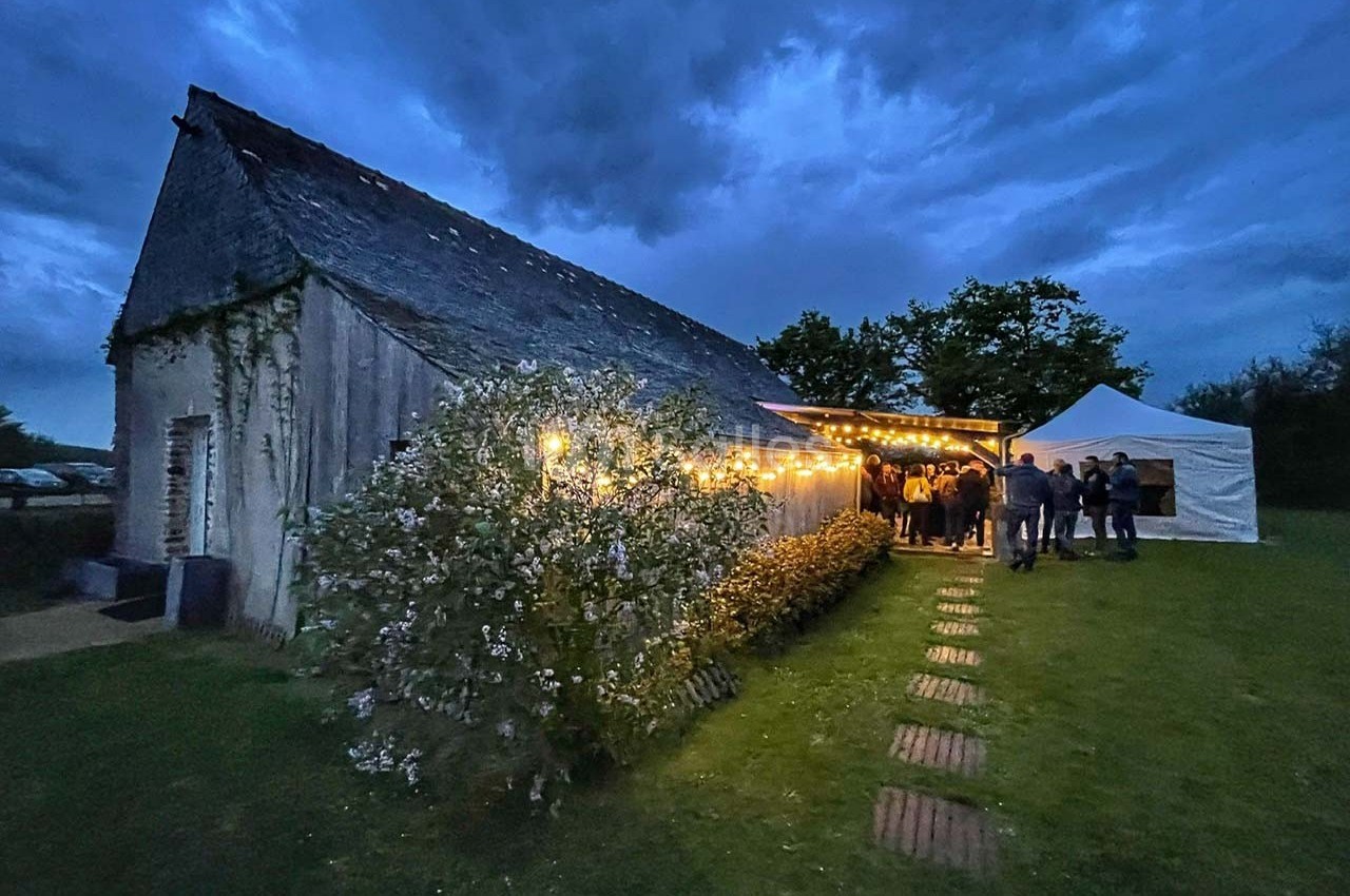 Bâtiment en pierre éclairé par des guirlandes lumineuses, avec des invités rassemblés sous un auvent au crépuscule.