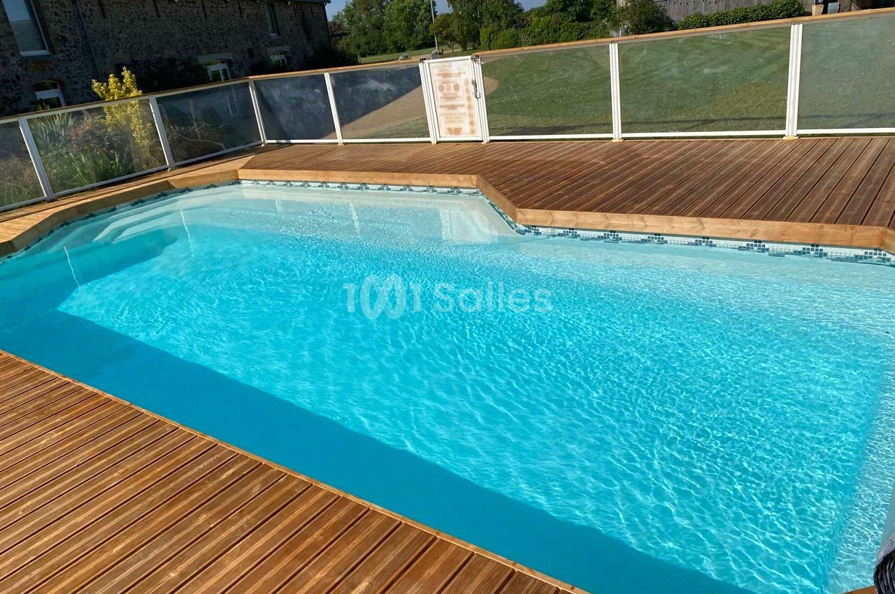Piscine extérieure entourée d'une terrasse en bois avec vue sur un jardin et des bâtiments en arrière-plan.