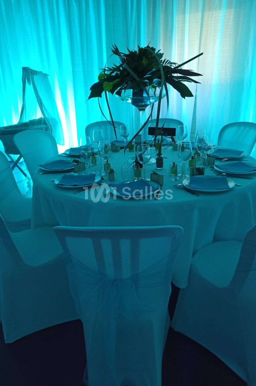 Table ronde décorée avec nappes blanches, vaisselle élégante et centre de table floral, éclairage bleu en arrière-plan.