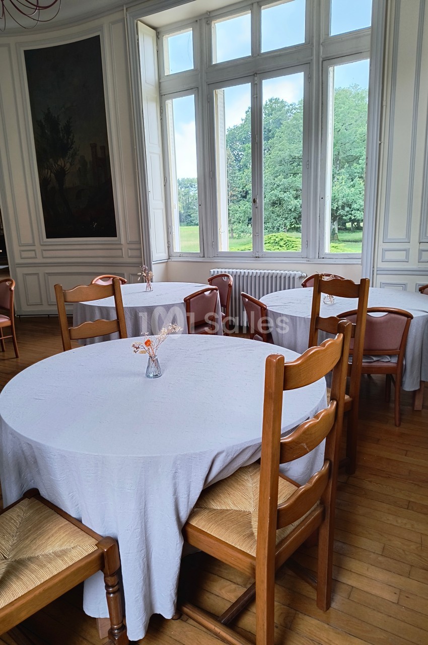 Salle lumineuse avec tables rondes couvertes de nappes blanches, chaises en bois et vue sur un jardin à travers de grandes…