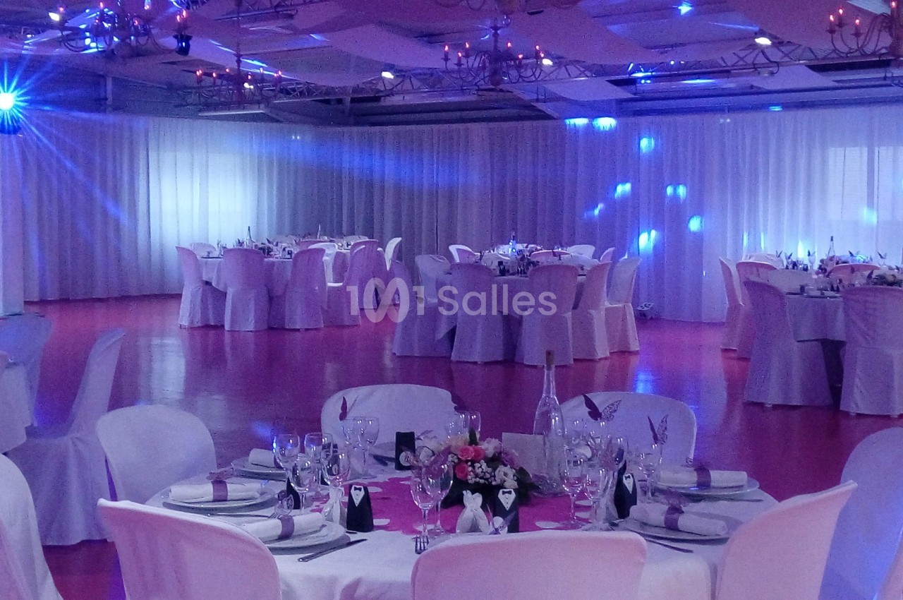 Salle de réception décorée avec des tables rondes dressées, nappes blanches et éclairage tamisé bleu.