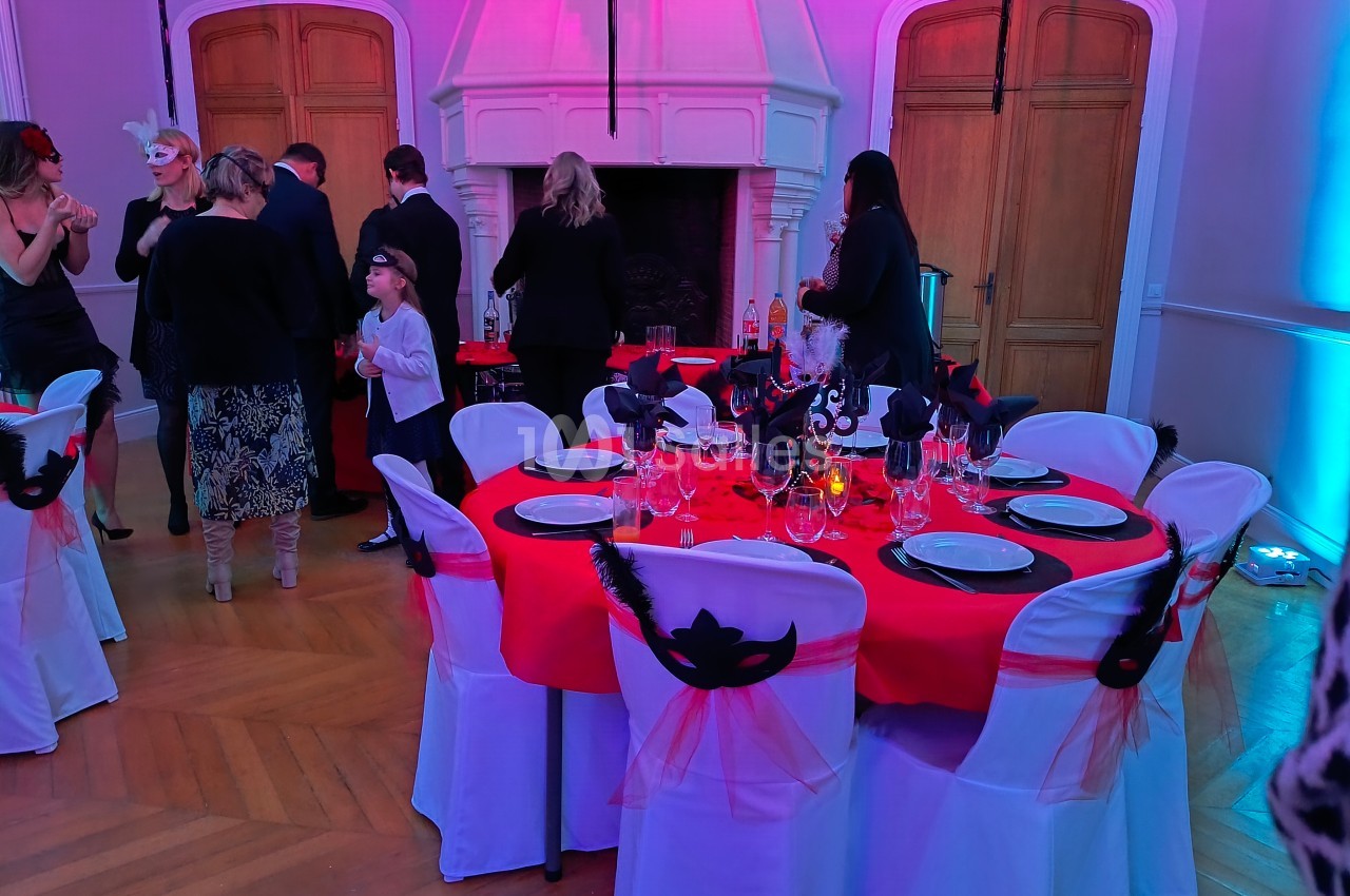 Salle décorée pour un événement, avec tables dressées en rouge et blanc, ornées de masques noirs et plumes.