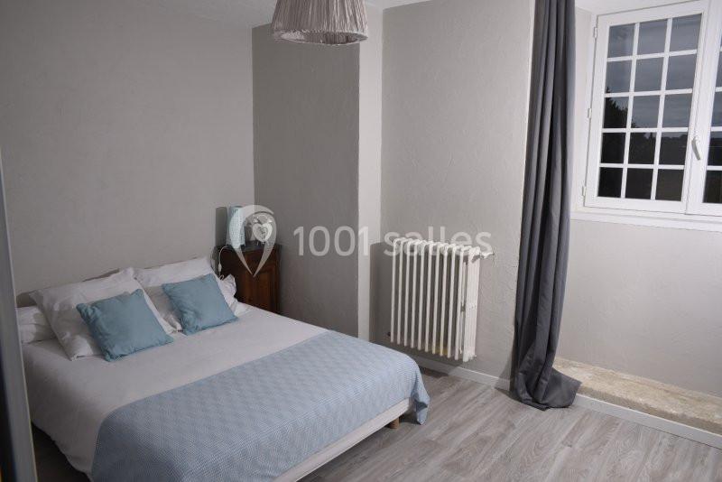 Chambre lumineuse avec lit double, coussins bleus, radiateur, fenêtre à carreaux et rideaux gris.