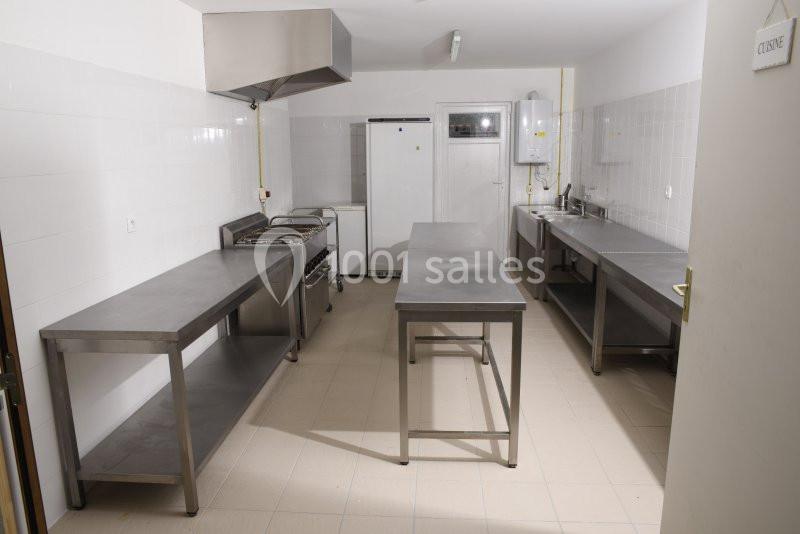 Cuisine professionnelle avec plans de travail en inox, cuisinière, réfrigérateur, évier et murs carrelés blancs.