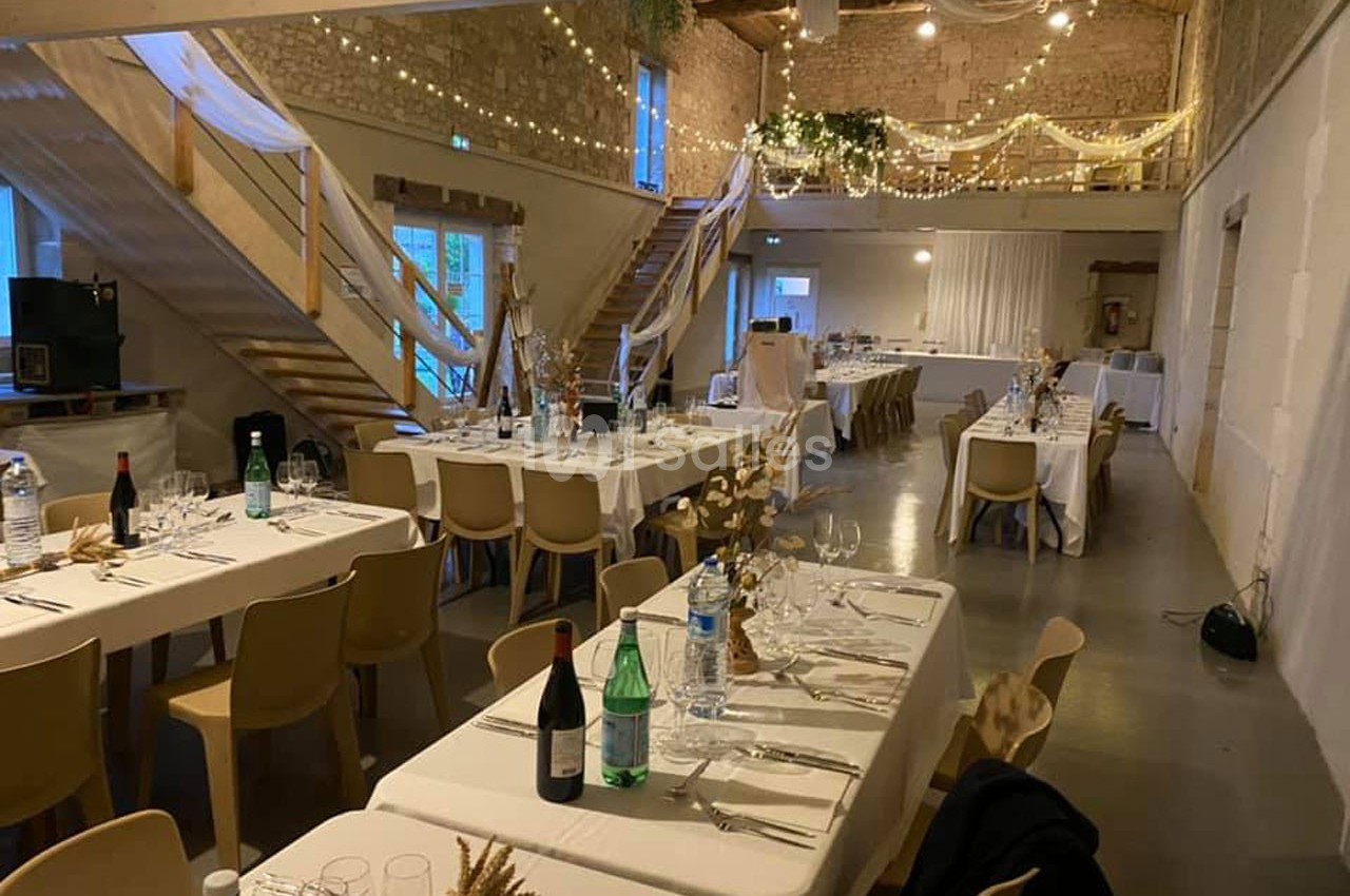 Salle de réception décorée avec des tables dressées, des guirlandes lumineuses et un plafond en bois apparent.