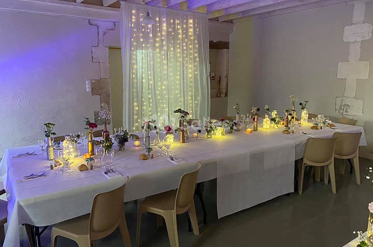 Grande table décorée pour un repas, ornée de fleurs, bougies et guirlandes lumineuses dans une salle au plafond en bois.