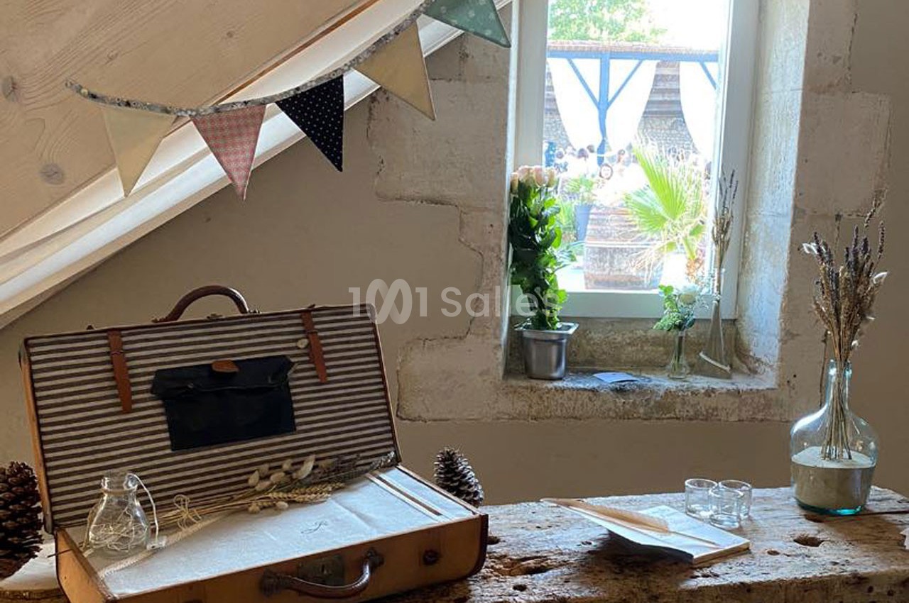 Valise ouverte sur une table en bois, décorée de fleurs séchées, sous une fenêtre avec guirlande de fanions.