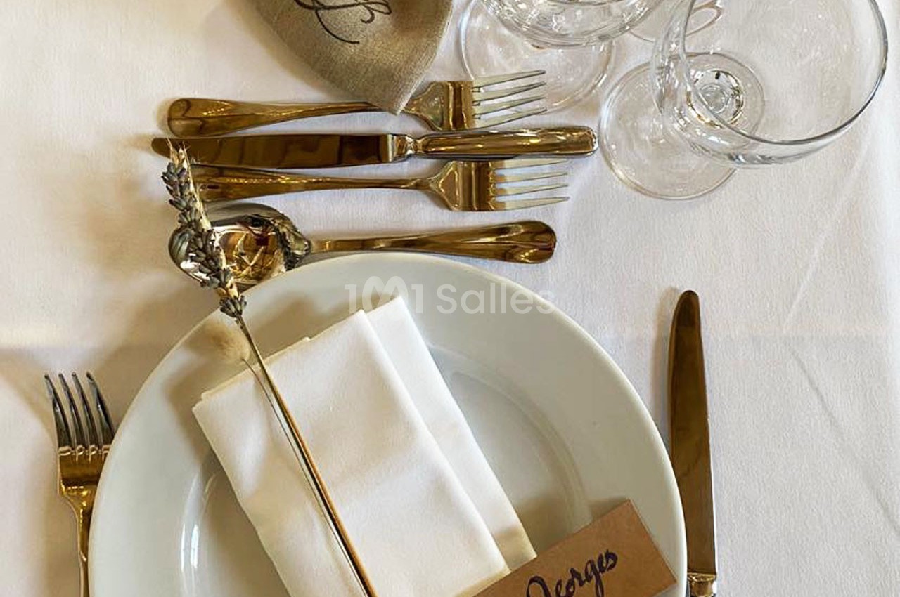 Table dressée avec assiette blanche, serviette pliée, couverts dorés, verres et étiquette portant un prénom.