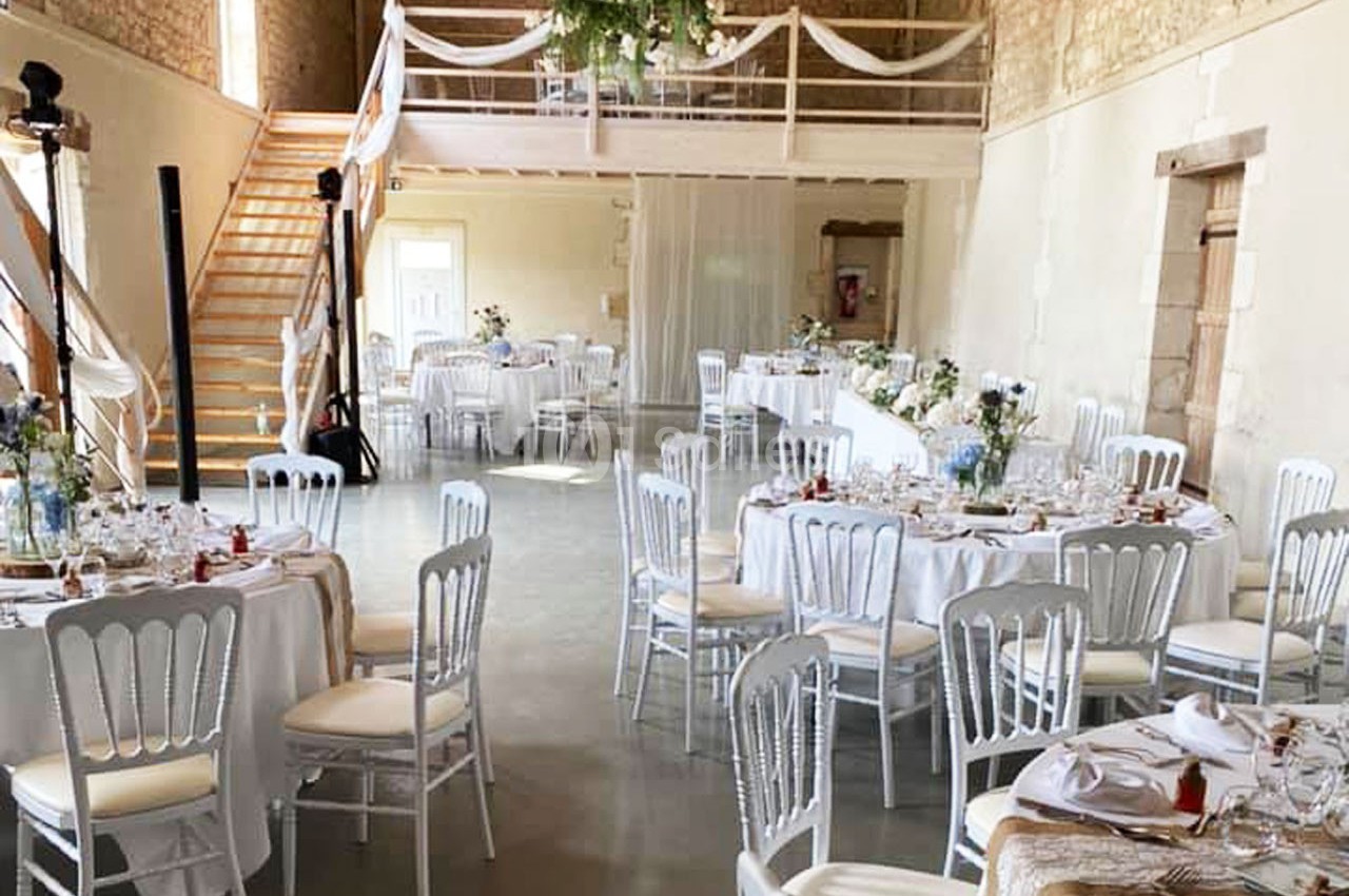 Salle de réception décorée avec des tables rondes dressées, des chaises blanches et des guirlandes suspendues.
