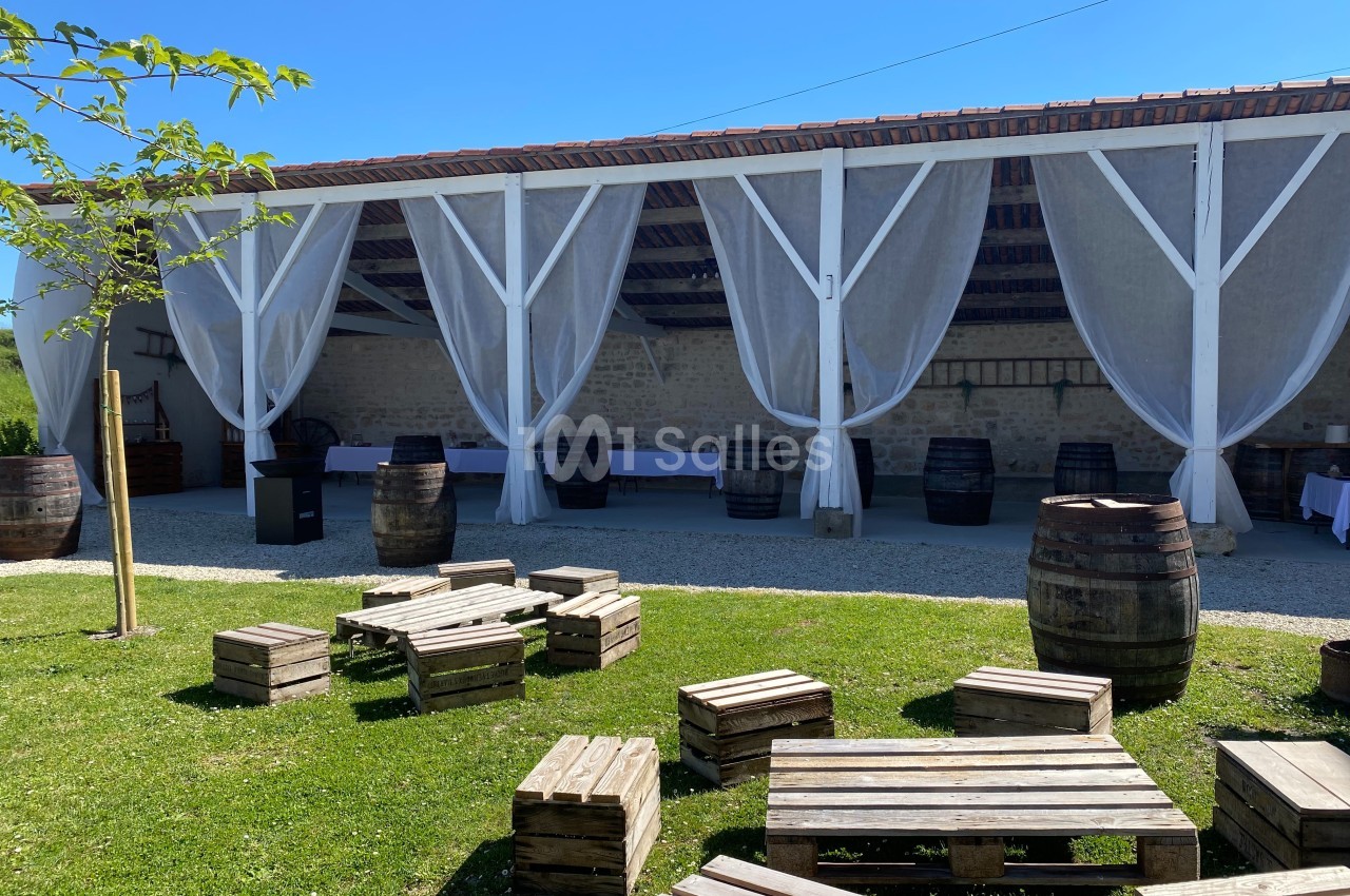 Terrasse couverte avec rideaux blancs, tonneaux en bois et palettes disposées sur une pelouse par une journée ensoleillée.
