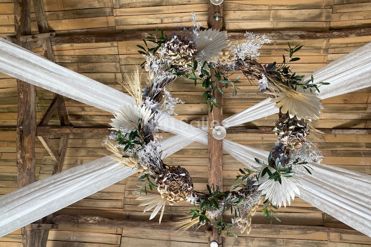 Couronne décorative suspendue sous un plafond en bois, ornée de feuillages, fleurs séchées et éléments naturels.