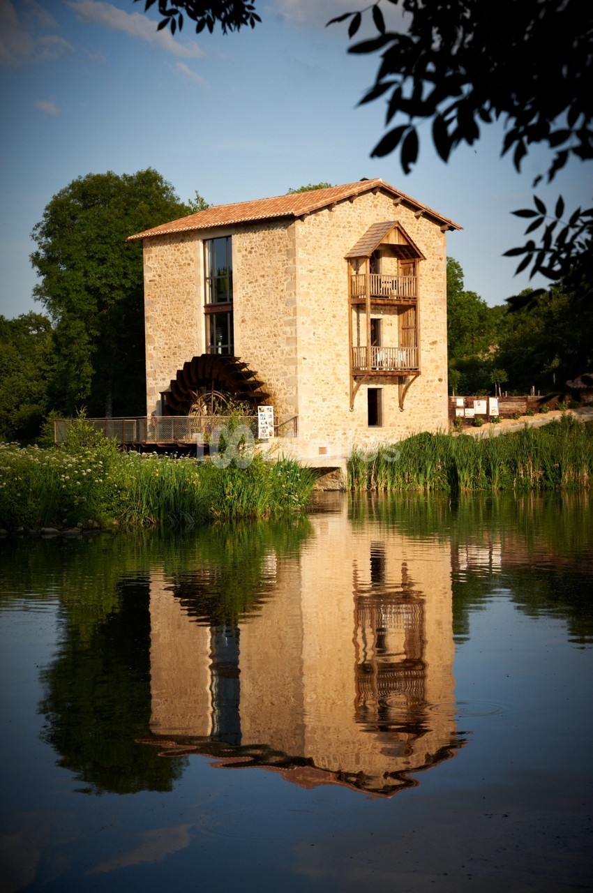 Bâtiment en pierre avec une roue à aubes, bordé par un plan d'eau et entouré de végétation.