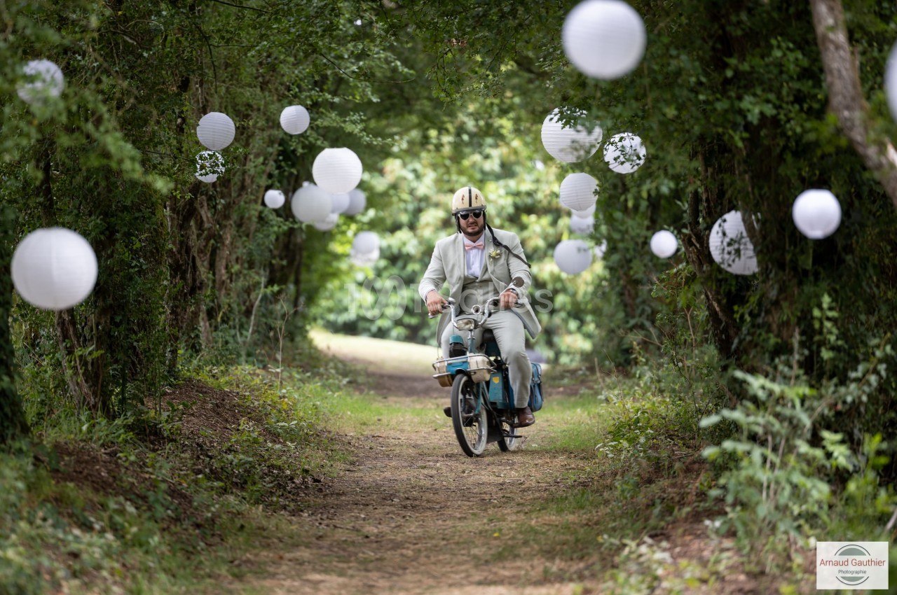Un homme en costume clair roule sur une mobylette dans un chemin forestier décoré de lanternes blanches suspendues.