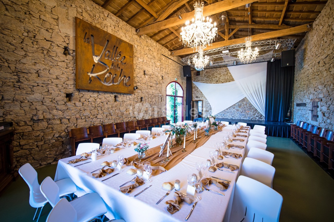 Salle en pierre avec une grande table dressée pour un repas, décorée de fleurs et éclairée par des lustres.