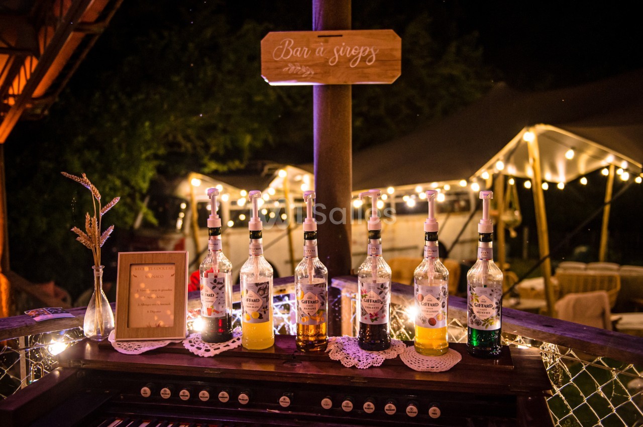 Bouteilles de sirops alignées sur un comptoir en bois, éclairées par des guirlandes lumineuses dans un cadre nocturne.