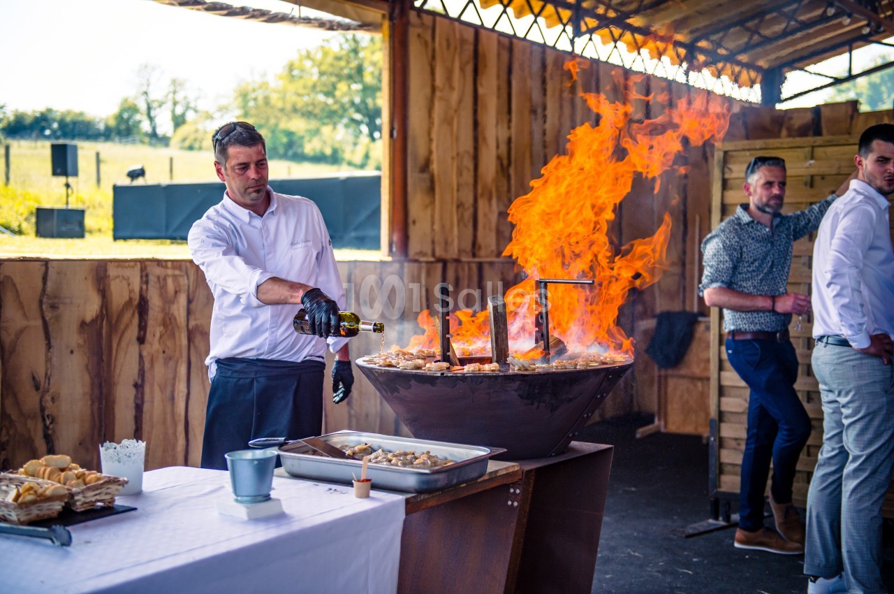 Un chef verse du liquide sur une grande plancha enflammée lors d'une cuisson en extérieur sous un abri en bois.